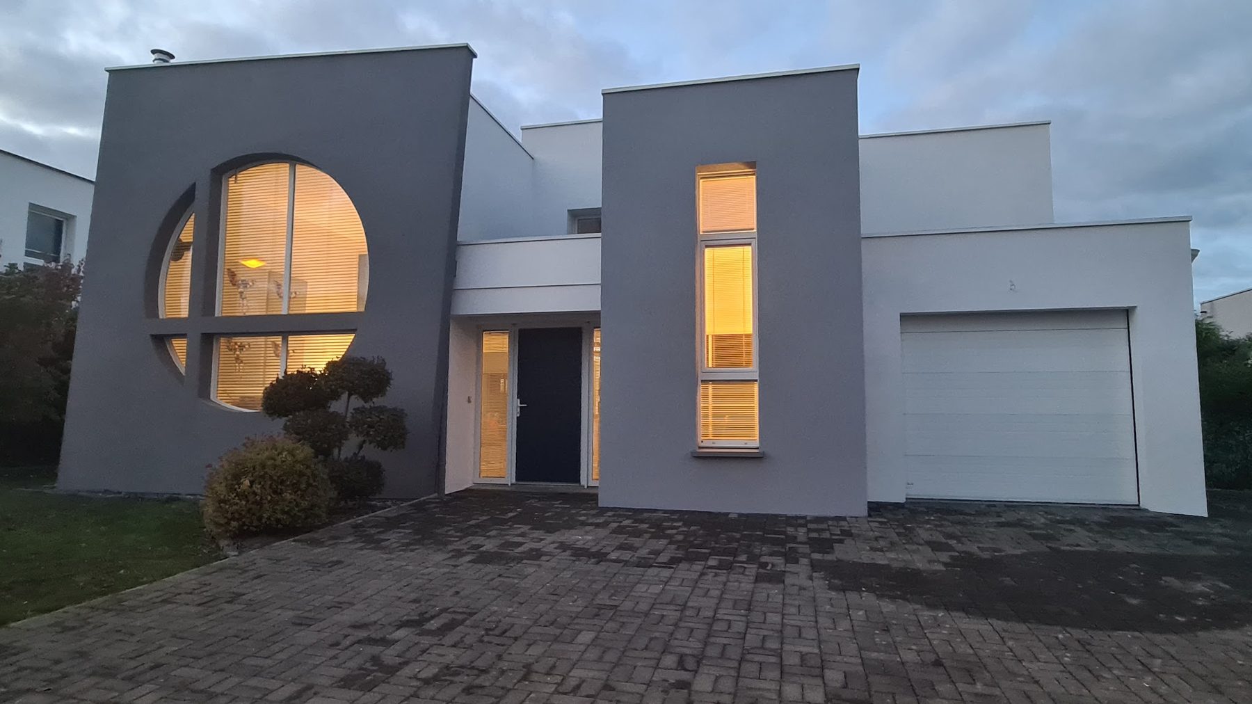 Maison contemporaine aux façades grise et blanche, fenêtre ronde éclairée, à la tombée du jour
